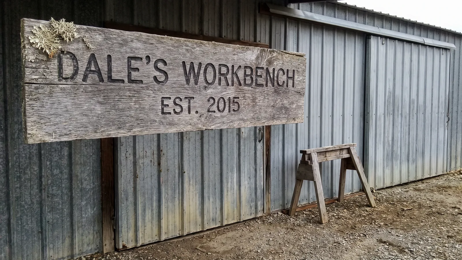 A weathered hand-carved wooden sign on the wall of a metal pole barn reading Dale's Workbench, Est. 2015